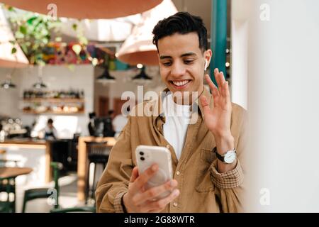 Lächelnder junger hispanischer Mann in Kopfhörern bei einem Videoanruf über Mobiltelefon, der im Café drinnen mit der Hand winkend steht Stockfoto