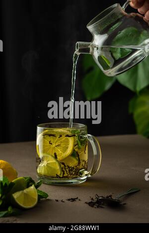 Frau gießt kochendes Wasser aus einer Teekane in einen Becher mit Minze, Zitrone, Limette und schwarzem Tee, Tee-Brauprozess Stockfoto