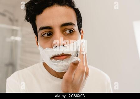 Junger hispanischer Mann, der sich sein Gesicht rasiert, stand im Innenbereich am Spiegel, aus der Nähe Stockfoto