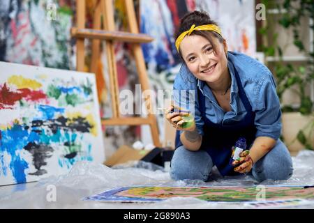 Glückliche junge Frau im Schürze lächelt der Kamera zu, hält den Pinsel während der Arbeit an der Malerei, sitzt auf dem Boden in der Werkstatt zu Hause Stockfoto