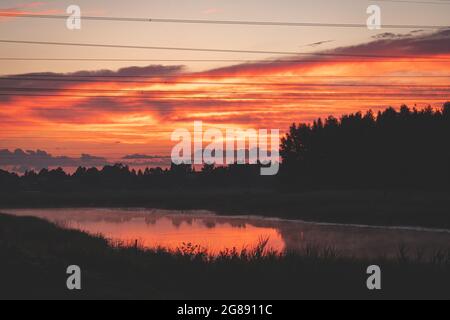 Sonnenuntergang über dem Fluss. Rot-orange Wolken. Schwarze Waldsilhouette über dem Fluss. Elektrische Stromleitungen in der Luft. Spiegelung von Wolken und Schilf Stockfoto