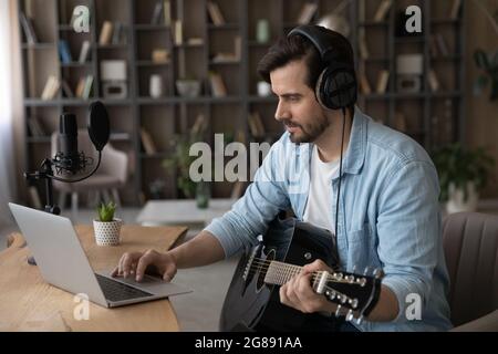 Glücklicher junger Mann spielt Gitarre mit Computer-Anwendung. Stockfoto
