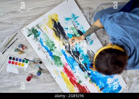 Draufsicht auf eine geschäftige junge Frau, eine Malerin, die an farbenfroher abstrakter Malerei arbeitet und in der heimischen Werkstatt auf dem Boden liegt Stockfoto