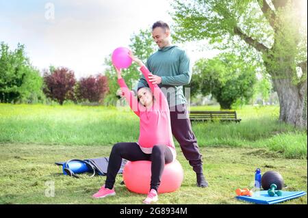 Lächelnder persönlicher Trainer und eine glückliche Schwangere, die Yoga-Übungen im Freien in einem Park macht - Girl does muscle streckt auf dem Gymnastikball sitzend - Konzept A Stockfoto