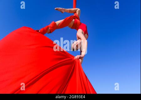 Outdoor-Aktivität der Turnerin, die Training auf roten Luftseiden und Bändern am Himmel durchführt - Mädchen, die Tanz in der Luft mit roten Stoffen, Sport, Stockfoto
