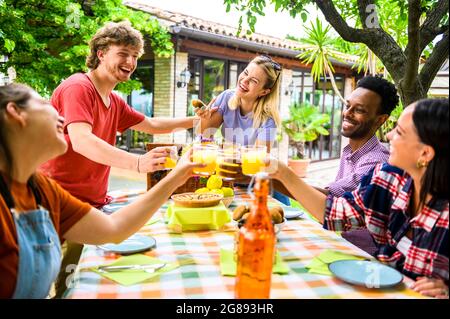 Junge multirassische Freunde, die Orangensaft auf der Gartenparty toasten - Millennial-Studenten-Gruppe, die Spaß beim gemeinsamen Frühstück im Sommer hat - Life Sty Stockfoto