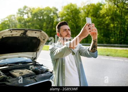 Trauriger, tausendjähriger Mann, der auf der Straße neben einem kaputten Auto steht, die Hand mit dem Handy hebt und versucht, den Pannendienst anzurufen Stockfoto