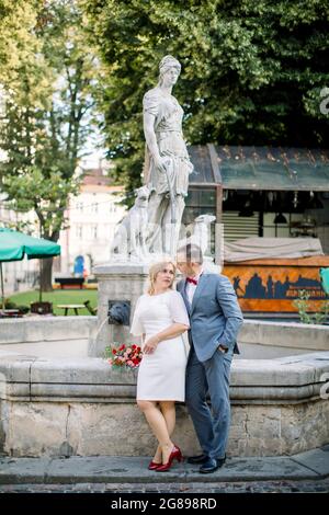 Liebende Paare mittleren Alters umarmen, während sie sich nach hinten auf den steinernen alten Stadtbrunnen lehnen und ihre Verabrege und ihren Spaziergang genießen. Schönes reifes Paar in der Altstadt Stockfoto