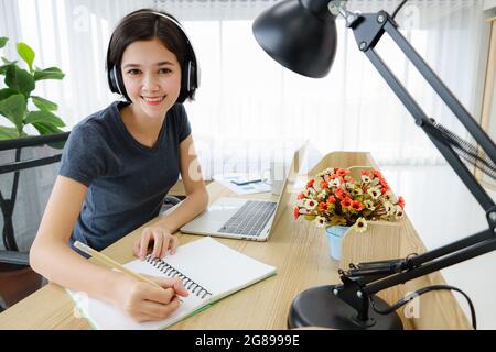 Junge, hübsche und wunderschöne Thai-deutsche Halbrennerin, die als Schreibtisch sitzt und lächelt, während sie im Schlafzimmer zu Hause arbeitet. Idee für Glück junge Menschen stu Stockfoto