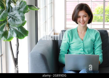 Junge, hübsche und wunderschöne Thai-deutsche Halbrennerin, die als Schreibtisch sitzt und lächelt, während sie im Wohnzimmer mit einem Labtop-Computer auf dem Sofa arbeitet. Idee für ha Stockfoto