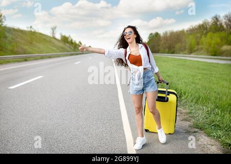 Fröhliche Millennial-Dame, die ihre Fahrt hinkitzeln lässt, Auto mit Koffer auf der Autobahn fängt, auf dem Land unterwegs ist, Platz kopieren Stockfoto