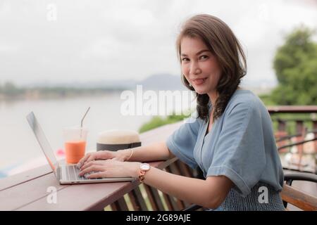 Schöne asiatische Frau mittleren Alters, die im Café neben dem Fluss sitzt und Kaffee trinkt und im Hintergrund die Berge sieht, während sie ein Notebook benutzt Stockfoto