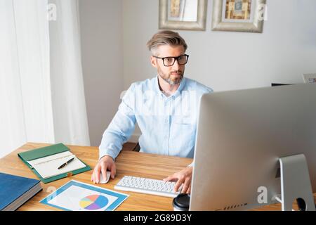 Geschäftsmann, der hinter seinem Computer sitzt, während er von zu Hause aus arbeitet. Stockfoto