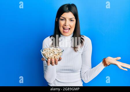 Junge hispanische Frau, die Kürbiskerne in der Schale hält und mit erhobener Hand Erfolge mit einem glücklichen Lächeln und einem Siegerausdruck feiert Stockfoto