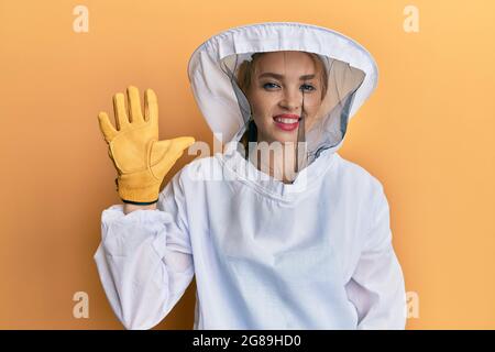 Schöne blonde kaukasische Frau in schützender Imkeruniform zeigt und zeigt mit den Fingern Nummer fünf nach oben, während sie selbstbewusst lächelt und hap Stockfoto