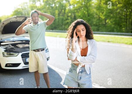 Junge gestresste Frau, die auf der Autobahn in der Nähe eines kaputten Autos steht, während ihr Freund Pannendienst auf dem Smartphone anruft Stockfoto