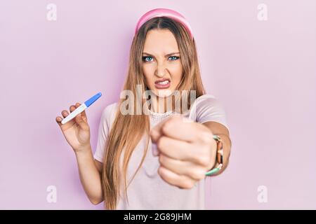 Junges blondes Mädchen, das das Ergebnis des Schwangerschaftstests hält, ist verärgert und frustriert und schreit vor Wut und erhobener Hand verrückt Stockfoto