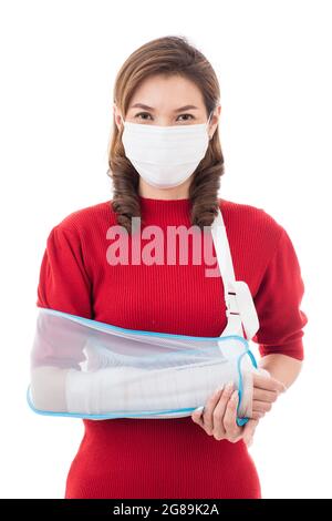 Schöne asiatische Frau mit gebrochenem Arm und Verband tragen chirurgische schützende Hygienemaske stehend isoliert auf weißem Hintergrund mit Smiley Gesicht. Ins Stockfoto