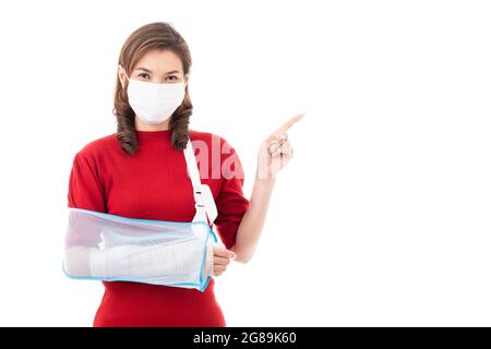 Schöne asiatische Frau mit gebrochenem Arm und Verband tragen chirurgische schützende Hygienemaske stehend isoliert auf weißem Hintergrund mit Smiley Gesicht. Poi Stockfoto