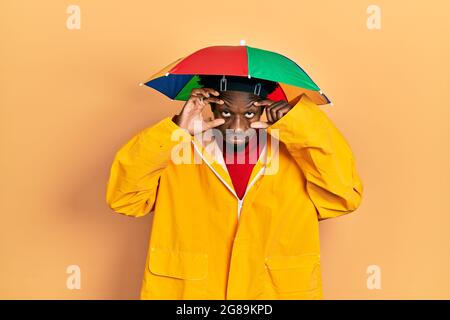 Junger afroamerikanischer Mann mit gelbem Regenmantel, der versucht, die Augen mit den Fingern zu öffnen, müde und müde wegen morgendlicher Müdigkeit Stockfoto