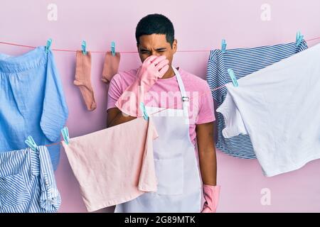 Junger gutaussehender hispanischer Mann mit sauberer Schürze, der Kleidung auf der Wäscheleine hält, die etwas stinkendes und ekelhaftes riecht, unerträglichen Geruch, hält Stockfoto