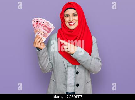 Schöne hispanische Frau mit islamischem Hijab, die chinesische Yuan-Banknoten hält, lächelt glücklich und zeigt mit Hand und Finger Stockfoto