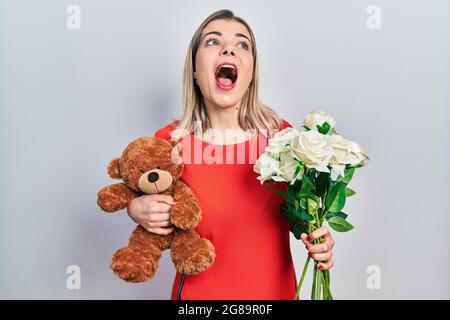 Schöne kaukasische Frau hält Bär und Blumenstrauß für Jahrestag wütend und verrückt Schreien frustriert und wütend, Schreien mit Wut Blick Stockfoto