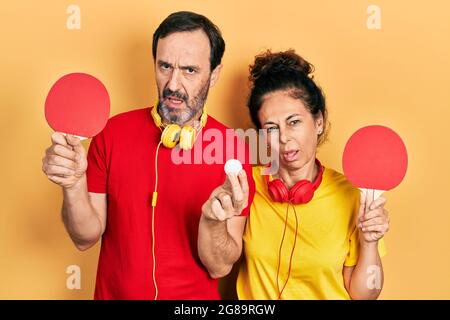 Ein Paar hispanischer Frauen und Männer mittleren Alters mit roten Tischtennisschlägern im Schockgesicht, die skeptisch und sarkastisch aussahen und mit offenem Mund überrascht waren Stockfoto