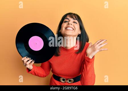 Junge hispanische Frau mit Vinylscheibe, die den Sieg mit einem glücklichen Lächeln und mit erhobenen Händen zum Ausdruck bringt Stockfoto