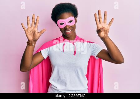 Junge afroamerikanische Mädchen trägt Superhelden-Maske und Cape-Kostüm zeigt und zeigt mit den Fingern Nummer neun, während sie selbstbewusst und glücklich lächelt Stockfoto