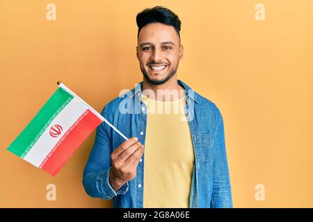 Junger araber, der die iranische Flagge hält, sieht positiv und glücklich aus und lächelt mit einem selbstbewussten Lächeln, das Zähne zeigt Stockfoto