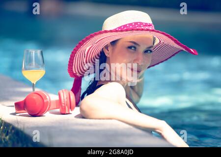 Eine asiatische Frau, die einen roten großen Hut trägt und im Pool mit einem Glas Orangensaft und Kopfhörern steht und mit einem glücklichen Gesicht auf die Kamera schaut. Stockfoto