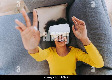 Schöne Frau, die zu Hause mit einem VR-Headset spielt - Schwarze Frau, die sich auf dem Sofa niederlegt, während sie mit der Augmented Reality-Technologie interagiert Stockfoto