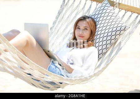 Schöne asiatische Frau trägt Sonnenbrille legte sich auf Hängematte in der Nähe des Meeres mit entspannenden und glücklichen Pose mit Laptop-Computer an sonnigen Tag mit blau Stockfoto