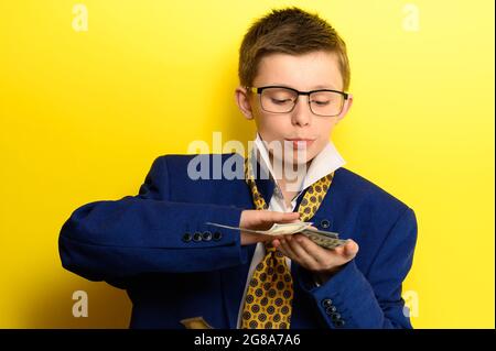 Junge in einem Anzug für Erwachsene auf gelbem Hintergrund, internationale Währung in den Händen eines Kindes, Ersparnisse für Kinder für die Zukunft. Stockfoto