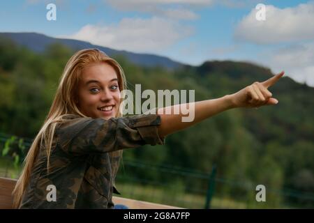 Aufgeregt motivierte junge blonde Frau, die mit weit geöffneten Augen in den Himmel zeigt und ein glückliches Lächeln im Freien in einer ländlichen Umgebung hat Stockfoto