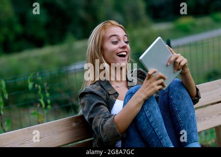 Fröhlich überraschte junge Frau, die ihr Tablet mit offenem Mund und weiten Augen anstarrt, während sie sich mit den Knien auf einer Bank im Garten entspannt Stockfoto