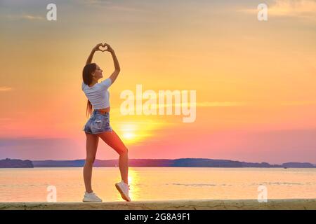 Junge Frau, die während des Sonnenuntergangs auf einem Pier steht und die Handflächen in Herzform zusammenfaltet Stockfoto