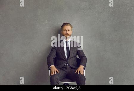 Serious selbstbewusst Geschäftsmann in eleganten Anzug und Krawatte sitzen auf einem Stuhl im Studio Stockfoto