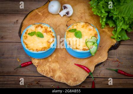 Töpfe mit Pilzjulienne auf einem Holzbrett, verziert mit Kräutern. Julienne in Kokotte mit gelber Käsekruste. Stockfoto