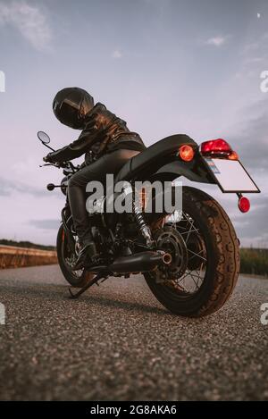 Stilvolle Motorradfahrerin Frau in Helm und Lederjacke sitzt auf Vintage-Motorrad. Weibliche Fahrerin im Freien auf Naturhintergrund. Ausflug, Cafe-Rennfahrer Stockfoto