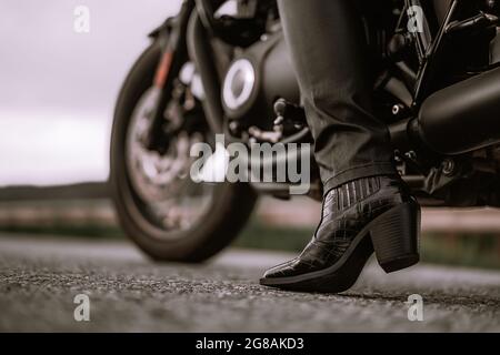Die Beine der stilvollen Motorradfahrerin Frau, die auf einem klassischen Fahrrad sitzt. Schwarzes Motorrad im Retro-Stil. Details des Vintage-Designs des brandneuen Motorrads. Stockfoto