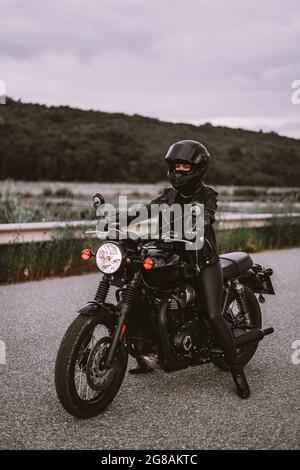 Stilvolle Motorradfahrerin Frau in Helm und Lederjacke sitzt auf Vintage-Motorrad. Weibliche Fahrerin im Freien auf Naturhintergrund. Ausflug, Cafe-Rennfahrer Stockfoto