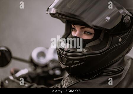 Stilvolle Motorradfahrerin Frau in Helm und Lederjacke sitzt auf schwarzem Motorrad. Weibliche Fahrerin im Freien auf Naturhintergrund. Ausflug, Cafe-Rennfahrer Stockfoto