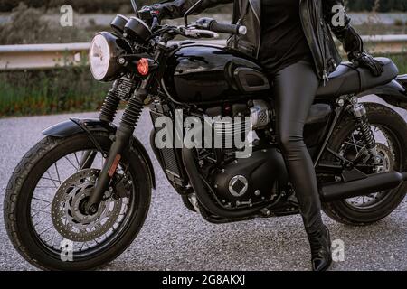 Die Beine der stilvollen Motorradfahrerin Frau, die auf einem klassischen Fahrrad sitzt. Schwarzes Motorrad im Retro-Stil. Details des Vintage-Designs des brandneuen Motorrads. Stockfoto