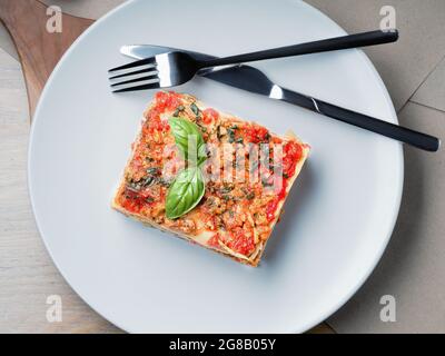 Lasagne. Weiße Platte mit Gabel und Messer. Basilikumblätter, Tomaten und Zucchini auf dem Tisch. Speisekarte mit italienischer Küche. Stockfoto