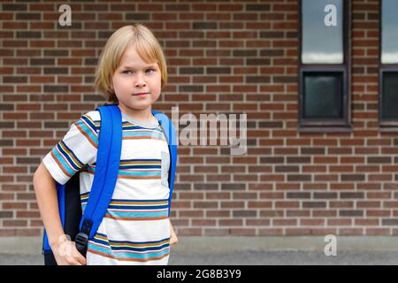 Niedliches Kind in der Nähe des Schulgebäudes auf dem Schulhof gegen eine Backsteinmauer mit Platz für Kopien. Kleiner blonder Schuljunge mit blauem Rucksack. Back to School Konz Stockfoto