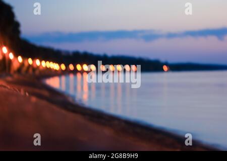 Verschwommene Abendpromenade am Meer in Unschärfe. Gelbe Laternen Lichter auf Laternenpfosten und blaues ruhiges Meer, Fluss in der Dämmerung. Die Nacht geht über eine Stadt. A stro Stockfoto