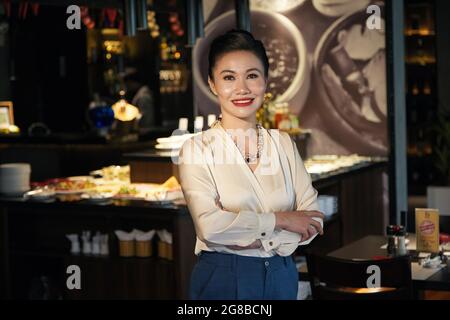 Porträt einer fröhlich lächelnden, eleganten Frau mittleren Alters, die an der Theke des Restaurants steht, das sie gerade eröffnet Stockfoto