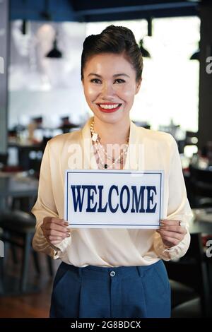 Porträt einer glücklichen, lächelnden Restaurantbesitzerin, die ein Begrüßungsschild in den Händen hält und die Kamera anschaut Stockfoto